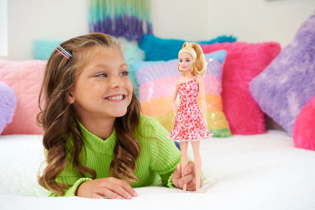 "Das Bild zeigt ein fröhliches Mädchen, das mit ihrer Barbie-Puppe spielt und dabei sichtlich Freude hat.
